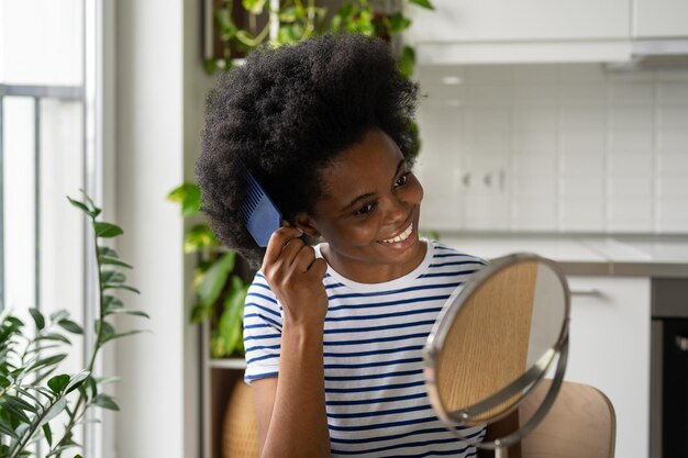 Une femme afro-américaine heureuse s'occupe des cheveux regarde dans le miroir avec un peigne en plastique se trouve dans l'appartement