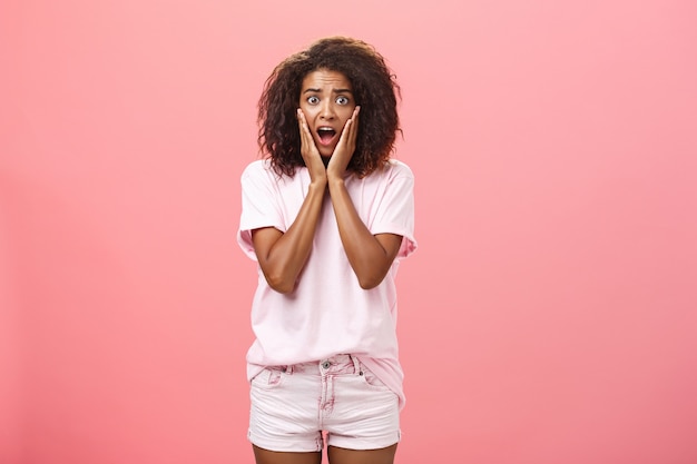 Femme afro-américaine inquiète avec une coiffure frisée en tenue à la mode avec la bouche ouverte tenant les mains sur le visage.