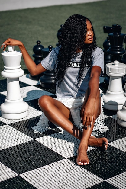 Femme afro-américaine intelligente jouant aux échecs géants alors qu'elle est assise sur le plateau avec les jambes croisées ; concept d'intelligence glamour.
