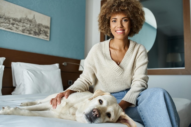 Une femme afro-américaine joyeuse câline son Labrador sur un lit dans une chambre d'hôtel adaptée aux animaux.