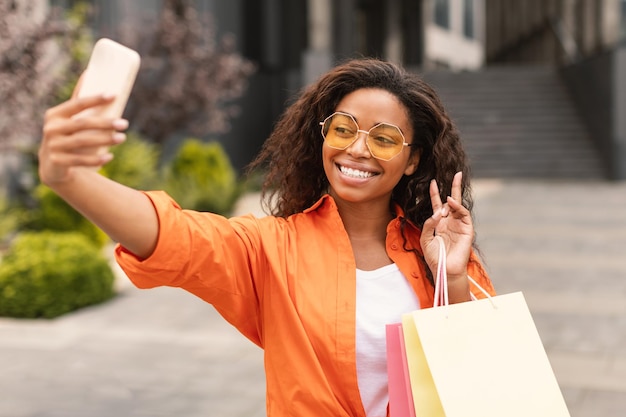 Une femme afro-américaine joyeuse du millénaire dans des verres avec de nombreux sacs avec des achats prend un selfie au téléphone