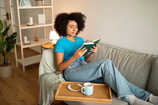 Une femme afro-américaine joyeuse du millénaire qui lit un livre savoure de la nourriture et boit du thé sur un canapé dans la vie