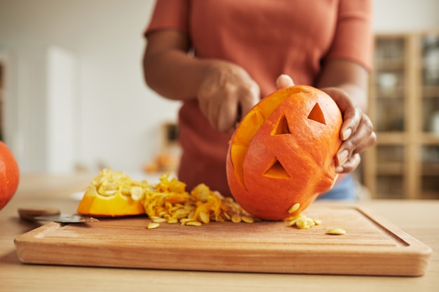 Femme afro-américaine méconnaissable debout à table sculpture Jack-O'-Lantern hors de citrouille orange mûre