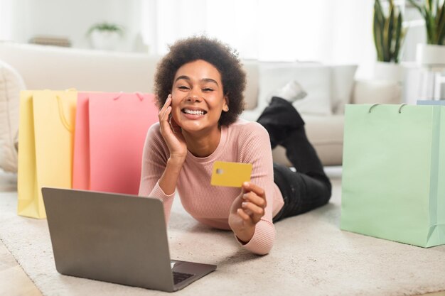 Photo une femme afro-américaine millénaire souriante avec de nombreux forfaits montre que la carte de crédit utilise un ordinateur portable se trouve sur le sol
