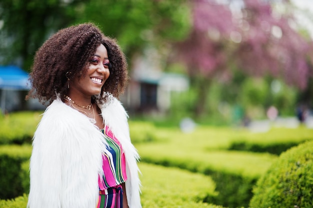 Femme afro-américaine à la mode en combinaison rayée rose avec manteau en fausse fourrure moelleux posé dans la rue de la floraison printanière