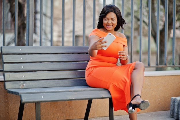 Femme afro-américaine modèle XXL en robe orange à la recherche sur téléphone mobile.