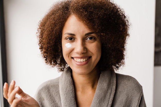 Une femme afro-américaine en peignoir applique une crème nettoyante et rajeunissante sur son visage