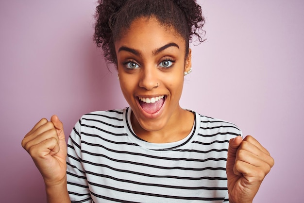 Femme afro-américaine portant un t-shirt rayé bleu marine debout sur un fond rose isolé criant fier et célébrant la victoire et le succès émotion d'encouragement très excitée