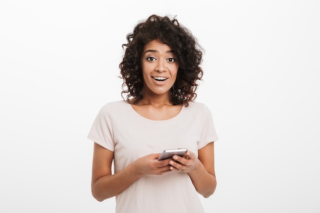 Femme Afro-américaine Positive Avec Des Cheveux Bouclés Tenant Un Téléphone Mobile Et, Isolé Sur Un Mur Blanc