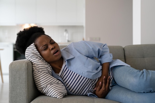 Une femme afro-américaine avec des problèmes de santé souffrant de crampes et de douleurs à l'estomac se trouve sur un canapé