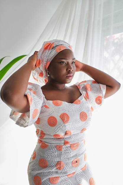 Femme afro-américaine réfléchie en tenue traditionnelle touchant la tête tout en se tenant près d'une fenêtre ouverte à l'arrière éclairée et en détournant le regard