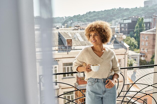 Photo une femme afro-américaine rêveuse debout avec une tasse de café sur le balcon de l'hôtel, vue sur la ville.