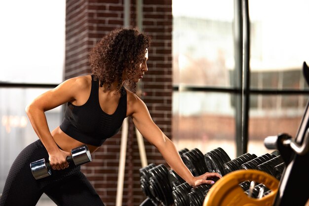 Une femme afro-américaine s'entraîne avec des haltères dans le gymnase devant un miroir.