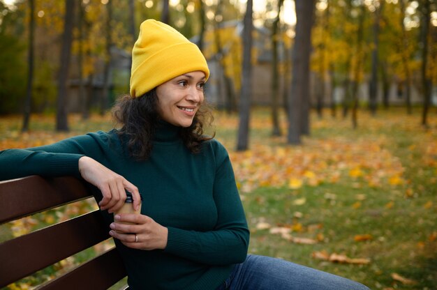 Une femme afro-américaine sereine et ravie vêtue de vêtements colorés et lumineux, assise sur un banc de parc avec une tasse à café en carton sur fond de chênes et d'érables d'automne, de jolis sourires regardant de côté