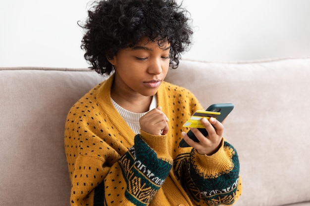 Femme afro-américaine shopping en ligne tenant un smartphone payant avec une carte de crédit or fille assise
