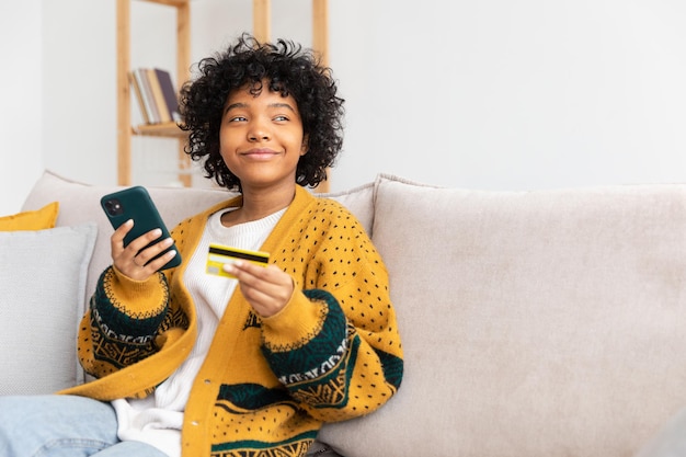 Femme afro-américaine shopping en ligne tenant un smartphone payant avec une carte de crédit or fille assise