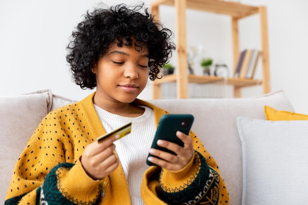 Femme afro-américaine shopping en ligne tenant un smartphone payant avec une carte de crédit or fille assise