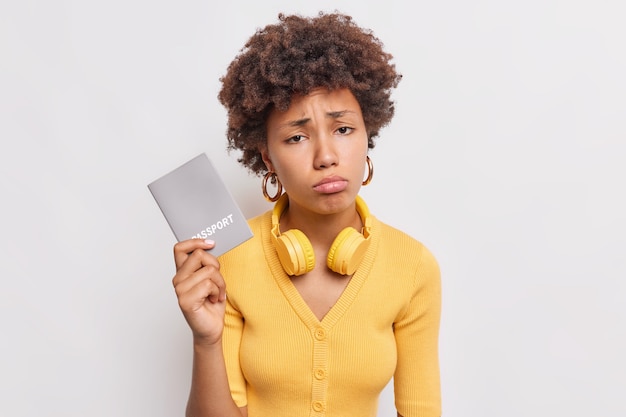 Une femme afro-américaine sombre et bouleversée aux cheveux bouclés tient un passeport se sent malheureuse car elle ne peut pas voyager pendant la pandémie de coronavirus porte des écouteurs sans fil autour du cou pose à l'intérieur