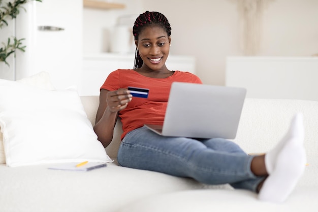 Une femme afro-américaine souriante effectuant des paiements en ligne avec un ordinateur portable et une carte de crédit