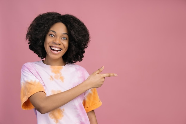 Femme afro-américaine souriante pointant le doigt sur l'espace de copie isolé sur fond rose Shopping