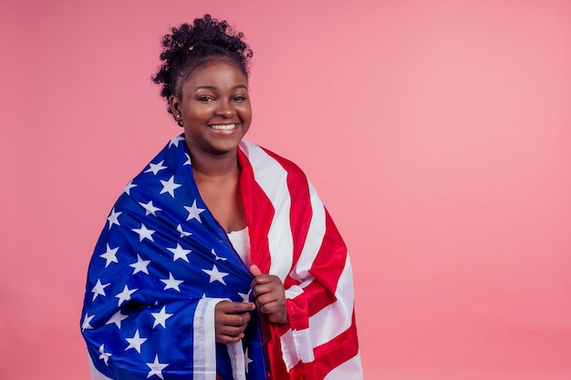 Femme afro-américaine souriante tenant le drapeau des États-Unis et regardant la caméra sur fond rose studio.