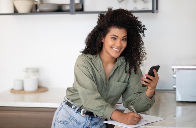 Femme afro-américaine souriante utilisant un téléphone portable prenant des notes dans la cuisine