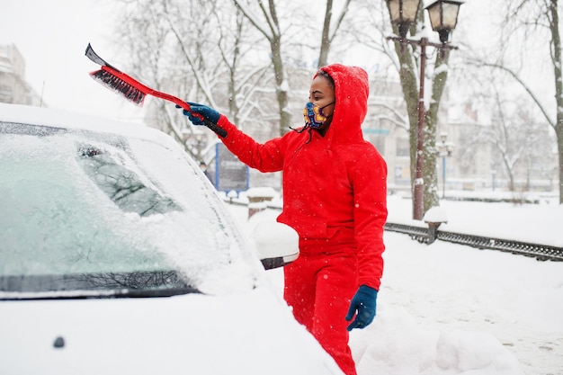 Une femme afro-américaine en sweat à capuche rouge et masque facial nettoie la voiture de la neige en hiver.