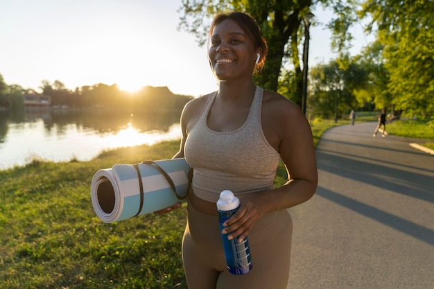 Une femme afro-américaine de taille plus grande se promène avec un tapis d'exercice et de l'eau dans le parc.