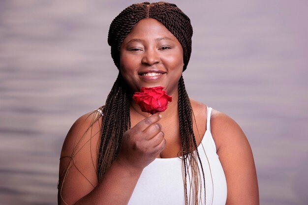 Femme afro-américaine tenant un portrait de symbole d'amour rose rouge. Dame heureuse souriante positive du corps avec fleur montrant le concept de beauté, de passion et de romance et regardant la caméra