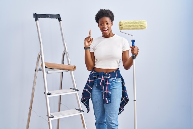 Femme afro-américaine tenant un rouleau peintre pointant le doigt vers le haut avec une idée réussie. excité et heureux. numéro un.