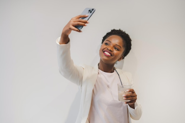 Photo femme afro-américaine tenant un verre de boisson et prenant un selfie avec son téléphone portable posant sur un mur blanc femme afro-américaine souriante tirant un selfie