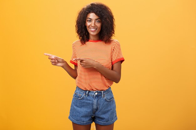 Femme afro-américaine en tenue décontractée pointant vers la gauche et souriant largement.