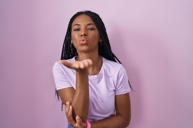 Femme afro-américaine avec des tresses debout sur fond rose regardant la caméra soufflant un baiser avec la main sur l'air étant une expression d'amour belle et sexy