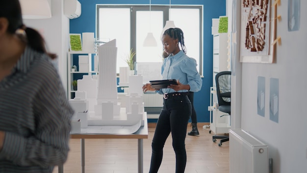 Femme afro-américaine utilisant une tablette numérique pour concevoir un plan de plans avec un modèle de construction sur table. Ingénieur industriel travaillant avec une maquette, planifiant l'aménagement et la structure de la construction.