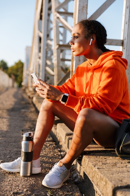 Photo femme afro-américaine en vêtements de sport utilisant des écouteurs et un téléphone portable assis sur le vieux pont