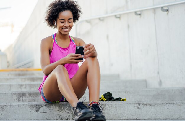 Femme afro athlétique à l'aide de son téléphone portable et se détendre après un entraînement à l'extérieur.