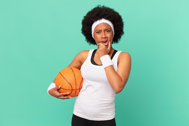 Femme afro avec la bouche et les yeux grands ouverts et la main sur le menton se sentant désagréablement choquée en disant quoi ou wow concept de basket-ball