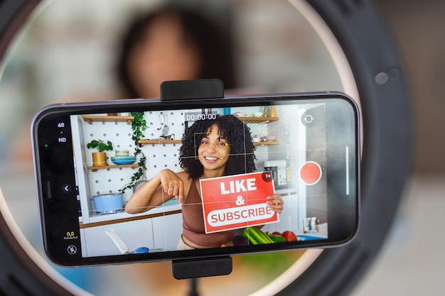 Une femme afro-caribéenne montrant une recette créative avec des ingrédients frais et sains de sa cuisine à la maison en utilisant son smartphone pour filmer la démonstration de cuisine