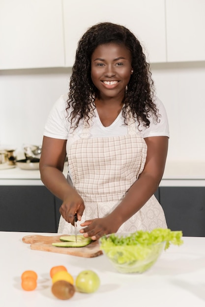 Femme afro coupant du concombre pour une salade fraîche jolie fille en tablier fait une salade pour le petit déjeuner près...