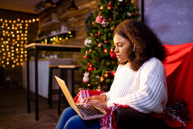 Femme afro faisant du shopping en ligne sur un ordinateur portable dans un intérieur de noël confortable