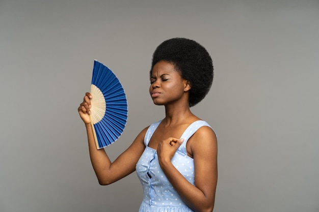 Une femme afro mécontente souffre de la chaleur à l'intérieur d'une femme africaine surchauffée agitant avec un ventilateur en papier