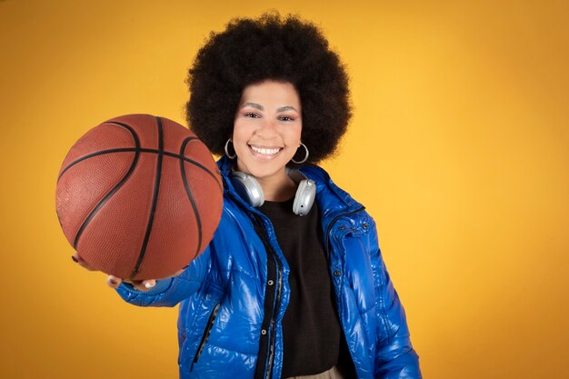 Femme afro mixte, portant des vêtements décontractés avec des écouteurs et un ballon de basket, isolé