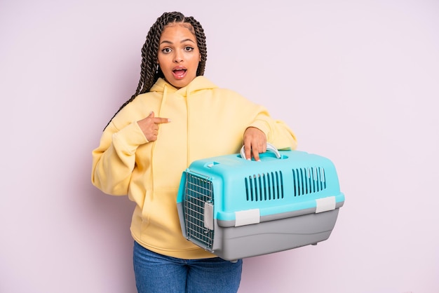 Femme afro noire à l'air choquée et surprise avec la bouche grande ouverte, pointant vers soi. concept de conteneur de voyage pour animaux de compagnie