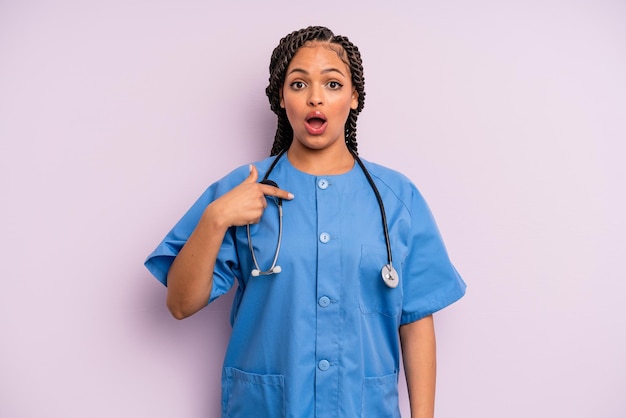 Femme afro noire semblant choquée et surprise avec la bouche grande ouverte pointant vers le concept d'auto-infirmière