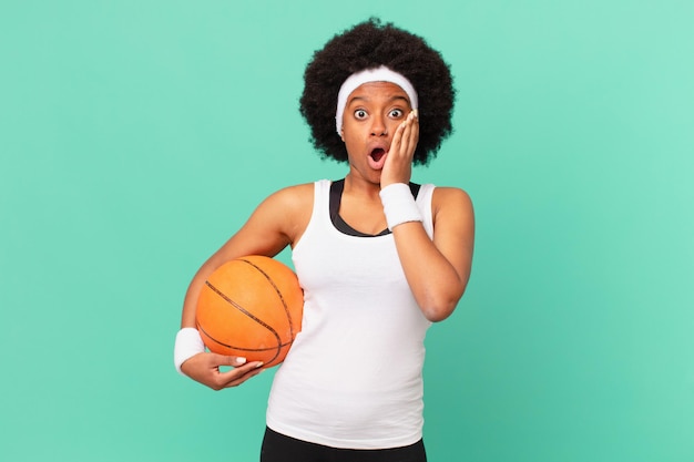 femme afro se sentant choquée et effrayée, l'air terrifiée avec la bouche ouverte et les mains sur les joues. concept de basket-ball