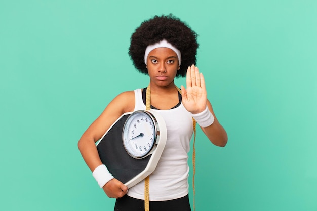 Femme afro semblant sérieuse, sévère, mécontente et en colère montrant la paume ouverte faisant un geste d'arrêt concept de régime