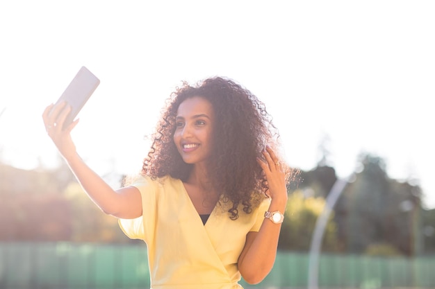 Une femme afro souriante aux cheveux bouclés prenant un selfie au coucher du soleil comprend un espace de copie et de texte