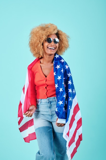 Femme afro en tenue décontractée et lunettes de soleil souriant tout en tenant un drapeau américain sur un fond isolé. Concept de la fête de l'indépendance des États-Unis et du patriotisme.