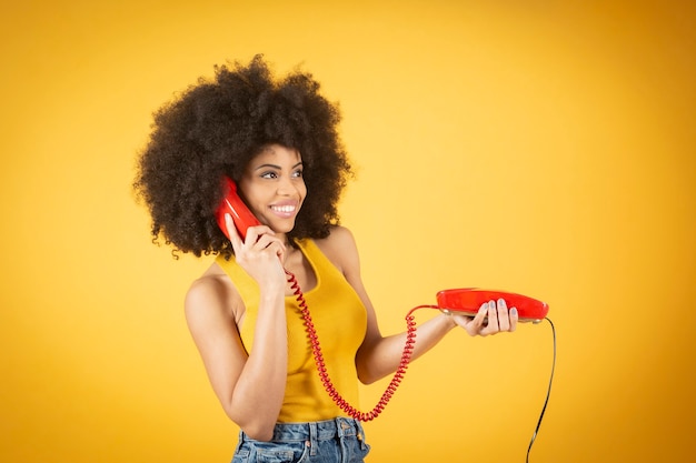 Femme afro avec un vieux téléphone par câble, fond jaune, téléphone rouge femme heureuse satisfaite des vêtements décontractés