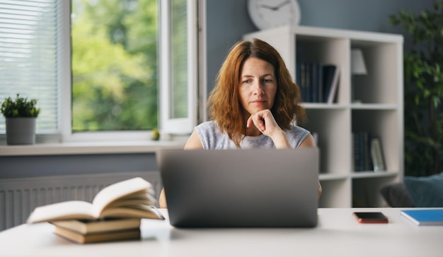 Femme d'âge moyen assise au bureau à domicile et regardant sur un écran d'ordinateur Belle femme travaillant sur un ordinateur portable sans fil dans un salon lumineux